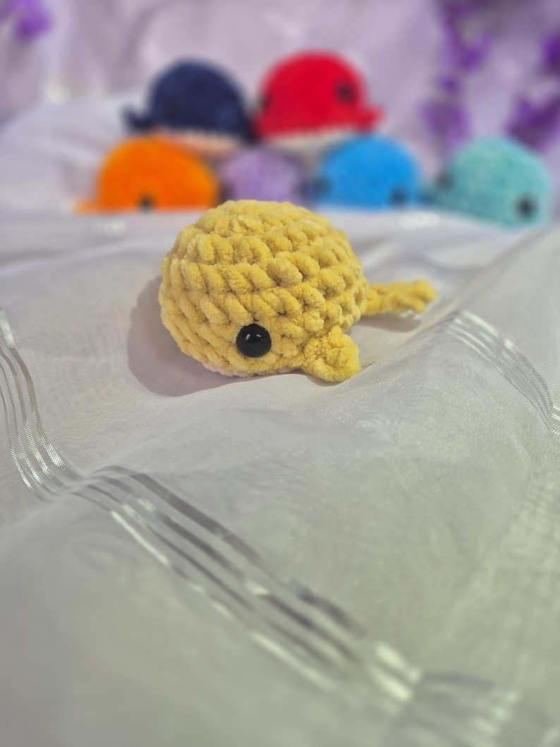 May include: A small, yellow, crocheted whale plushie made from soft, chunky yarn. The whale features a rounded body, a small tail, and a single visible black plastic eye on its side. Several other colourful crocheted whale plushies are blurred in the background.