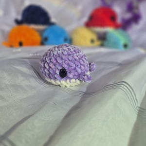 May include: A small, round, purple crocheted whale plushie with a white underbelly and black plastic eyes. The textured yarn creates a soft appearance. Several other colourful crocheted whale plushies are visible in the blurred background.