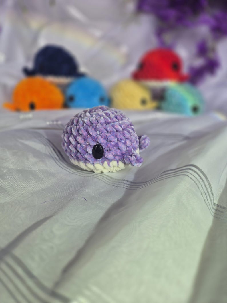 May include: A small, round, purple crocheted whale plushie with a white underbelly and black plastic eyes. The textured yarn creates a soft appearance. Several other colourful crocheted whale plushies are visible in the blurred background.