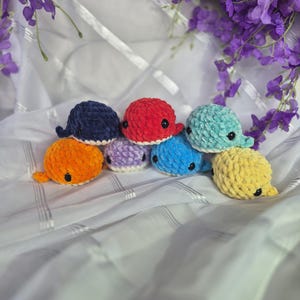 May include: A collection of seven small, plush, crocheted whale toys in various colours including navy blue, bright red, light teal, orange, light purple, sky blue, and yellow. Each whale features a soft, textured yarn finish and black eyes.