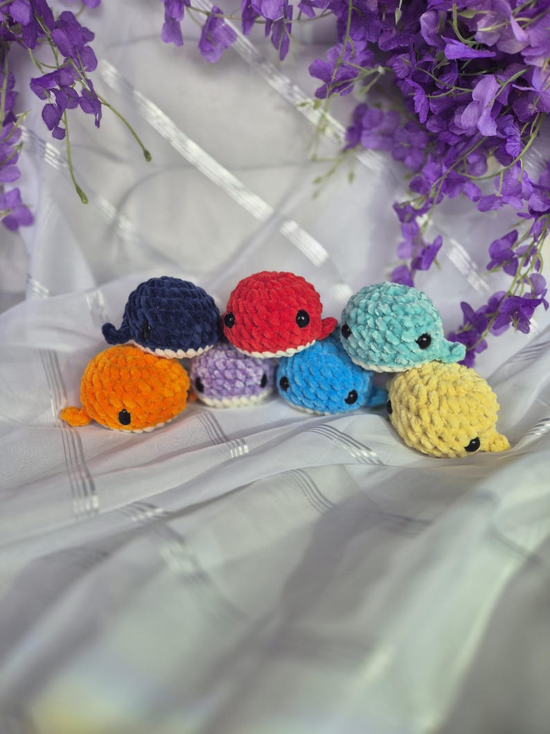 May include: A collection of seven small, plush, crocheted whale toys in various colours including navy blue, bright red, light teal, orange, light purple, sky blue, and yellow. Each whale features a soft, textured yarn finish and black eyes.