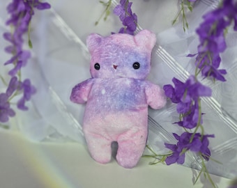 Könnte beinhalten: Ein kleiner Plüschbär mit einem weichen Batikmuster in Pastellrosa, Lavendel und Blau. Der Bär hat zwei kleine, runde schwarze Augen und eine winzige, aufgestickte rote Nase. Die Textur wirkt flauschig und weich, mit einem runden Kopf und Ohren.