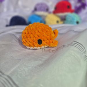 May include: A small, orange, crocheted whale plushie with a white underbelly and a black plastic eye. The whale has a rounded body and small fins. In the background, several other crocheted whale plushies in various colours are visible.