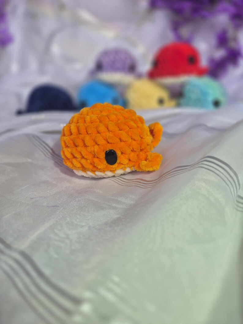 May include: A small, orange, crocheted whale plushie with a white underbelly and a black plastic eye. The whale has a rounded body and small fins. In the background, several other crocheted whale plushies in various colours are visible.