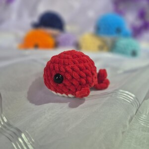 May include: A small, round, red crocheted whale plush toy with a textured yarn surface, a single black plastic eye, and a white underside. In the blurred background, several other similar crocheted whale plush toys in various colours are visible.