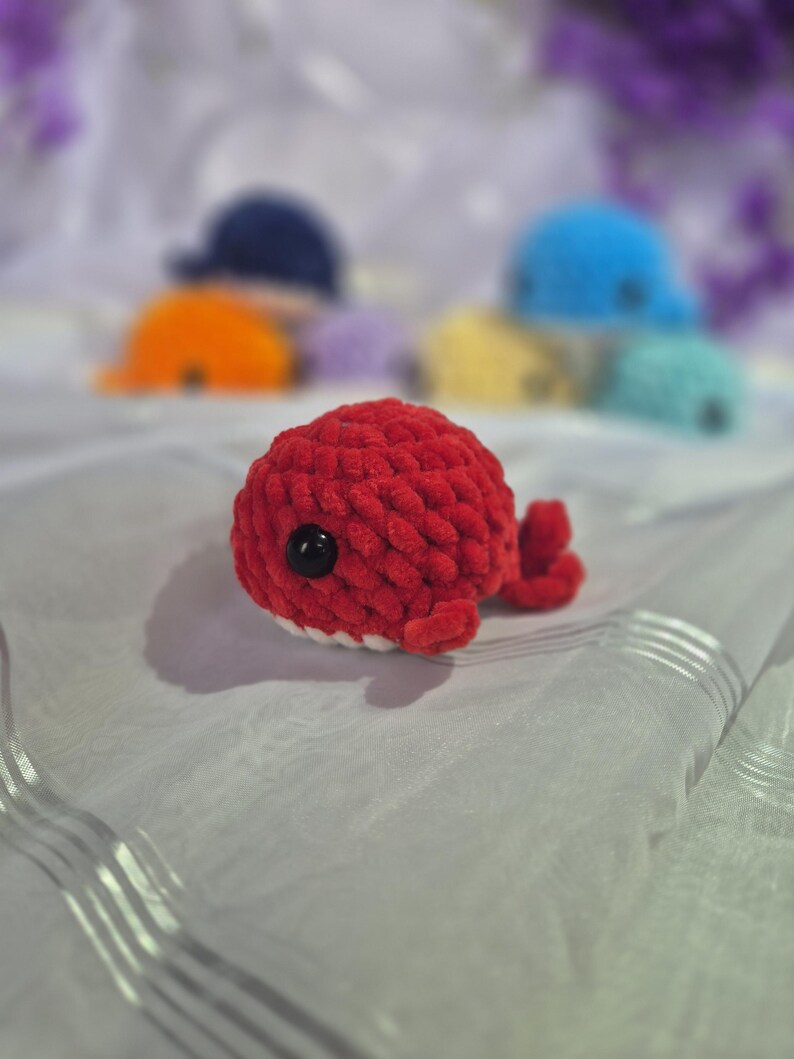 May include: A small, round, red crocheted whale plush toy with a textured yarn surface, a single black plastic eye, and a white underside. In the blurred background, several other similar crocheted whale plush toys in various colours are visible.