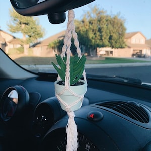 May include: A white macrame plant hanger with a small green succulent plant in a light blue pot. The hanger is attached to the rearview mirror of a car.