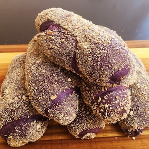 May include: A stack of purple sweet potato bread rolls, coated in a layer of breadcrumbs. The rolls are arranged in a pyramid shape on a wooden cutting board.