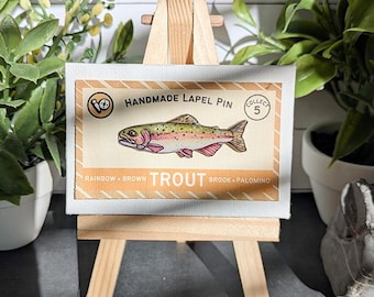 May include: A rectangular display card featuring a central illustration of a rainbow trout. The text reads HANDMADE LAPEL PIN, COLLECT 5, and RAINBOW BROWN TROUT BROOK PALOMINO. The card is mounted on a small wooden easel for display purposes.
