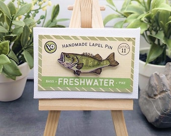 May include: A handmade lapel pin featuring a green bass fish illustration on a rectangular display card. The card has a light yellow background with green striped borders, text reading "HANDMADE LAPEL PIN", "FRESHWATER", "BASS", "PIKE", and "COLLECT 11".