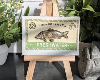 May include: A rectangular display card featuring a detailed illustration of a carp fish. The card is labeled HANDMADE LAPEL PIN and FRESHWATER, with the text COLLECT 11 in a circle. The card is mounted on a small wooden easel for display.