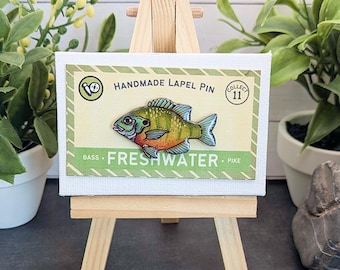 May include: A lapel pin shaped like a colorful fish with green, yellow, and orange scales, mounted on a rectangular card labeled HANDMADE LAPEL PIN, FRESHWATER, BASS, and PIKE, displayed on a small wooden easel stand.