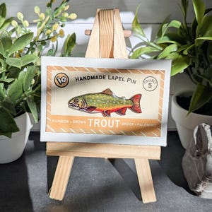 May include: A rectangular display card featuring a colorful illustration of a trout fish. The text reads Handmade Lapel Pin, Collect 5, and Rainbow, Brown, Trout, Brook, Palomino. The card is mounted on a small wooden easel for display purposes.