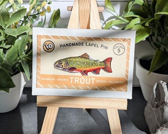 May include: A rectangular display card featuring a colorful illustration of a trout fish. The text reads Handmade Lapel Pin, Collect 5, and Rainbow, Brown, Trout, Brook, Palomino. The card is mounted on a small wooden easel for display purposes.