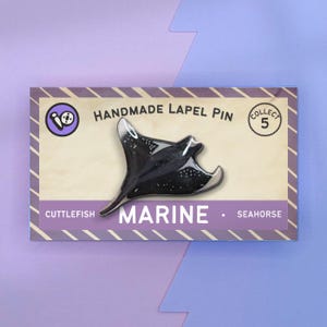 May include: A black and white enamel pin of a manta ray on a card with the text "HANDMADE LAPEL PIN" and "MARINE". The card also has a small circle with the number "5" inside and the text "COLLECT".