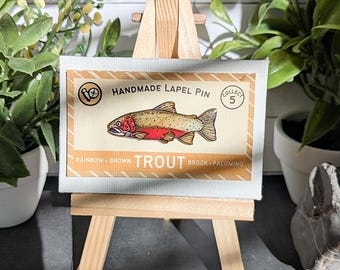 May include: A rectangular display card featuring a graphic illustration of a rainbow trout with a red stripe, set against a tan background. The text reads HANDMADE LAPEL PIN, COLLECT 5, and RAINBOW BROWN TROUT BROOK PALOMINO. The card is mounted on a wooden easel.