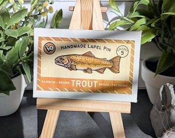 May include: A rectangular display card featuring a brown trout illustration, the text "HANDMADE LAPEL PIN", "COLLECT 5", and "RAINBOW BROWN TROUT BROOK PALOMINO" on a tan background, resting on a small wooden easel. The card has a striped border.