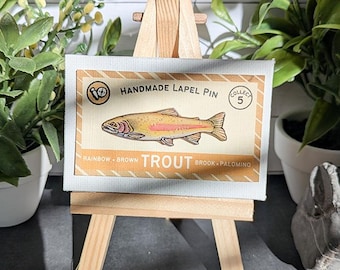 May include: A rectangular card featuring a graphic illustration of a rainbow trout fish centered on a tan background. The text reads HANDMADE LAPEL PIN, COLLECT 5, and RAINBOW BROWN TROUT BROOK PALOMINO. The card is displayed on a small wooden easel.