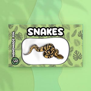 May include: A yellow and black snake enamel pin with the word "SNAKES" in a white bubble above it. The pin is on a green background with a tropical leaf pattern. The text "HANDMADE PIN" is on the left side of the pin.