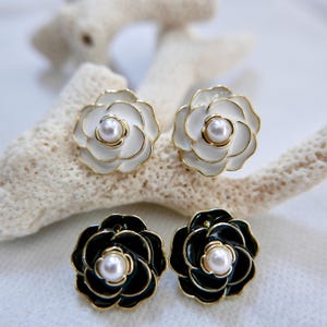 May include: A pair of earrings featuring a black and white flower design with a pearl in the centre. The earrings are gold-toned and have a classic, elegant style.