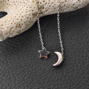 Puede incluir: Un par de pendientes de plata con un dije de estrella y media luna. Los pendientes están sobre una superficie blanca de tipo coral.