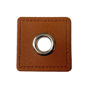Könnte beinhalten: Ein quadratischer, brauner Lederflicken mit einer silbernen Öse in der Mitte. Der Flicken hat einen genähten Rand. Das Leder hat ein strukturiertes Aussehen.