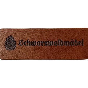 Könnte beinhalten: Braunes Leder-Etikett mit einer schwarzen Tannenzapfen und dem Text "Schwarzwaldmädel" in Schwarz.