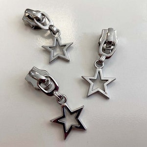 Könnte beinhalten: Drei silberne Reißverschluss-Zipper mit sternförmigen Anhängern. Die Sterne haben ein offenes Design und sind mit einer kleinen Schlaufe an den Reißverschluss-Zipper befestigt.