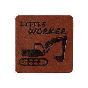 Könnte beinhalten: Ein brauner quadratischer Untersetzer mit den Worten "LITTLE WORKER" und einer schwarzen Bagger-Grafik. Der Untersetzer besteht aus einem strukturierten Material und ist vor weißem Hintergrund.