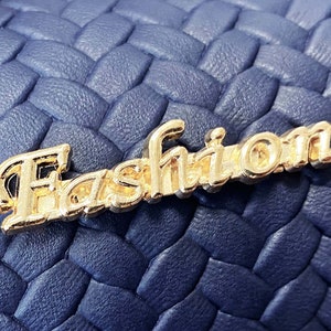 Op de afbeelding: Een close-up van een blauwe geweven stof met een gouden metalen "Fashion" label.