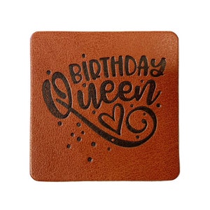 Könnte beinhalten: Quadratischer brauner Untersetzer mit den Worten "BIRTHDAY Queen" in schwarzer Schrift. Unter dem Wort "Queen" befindet sich ein Herzdesign. Der Untersetzer hat ein strukturiertes Aussehen und ist wahrscheinlich aus Leder oder einem ähnlichen Material.