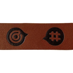 Könnte beinhalten: Ein rechteckiger brauner Lederaufnäher mit zwei schwarzen Sprechblasen-Symbolen. Das linke Symbol ist ein Zielsymbol, das rechte ein Hashtag-Symbol. Der Aufnäher hat eine strukturierte Oberfläche und ein schlichtes Design.