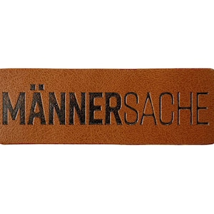 Könnte beinhalten: Ein rechteckiges, braunes Lederetikett mit dem schwarzen Schriftzug "MÄNNERSACHE". Das Etikett hat abgerundete Ecken und eine strukturierte Oberfläche, vor weißem Hintergrund.