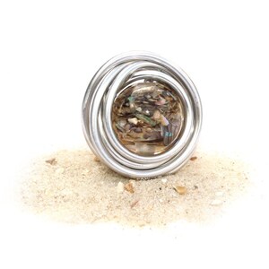May include: A silver wire ring with a round, clear glass orb filled with iridescent seashells. The ring is sitting on a pile of sand.