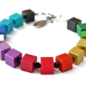 Puede incluir: Un brazalete de colores del arcoíris hecho de cuentas cuadradas con acentos negros. El brazalete está hecho de una variedad de colores, incluyendo rojo, naranja, amarillo, verde, azul y morado.