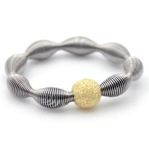 Könnte beinhalten: Ein silberner Ring mit einer strukturierten goldenen Perle in der Mitte. Der Ring besteht aus mehreren gewickelten Metallteilen.