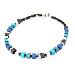 Può includere: Una collana realizzata con perline quadrate blu, turchese, bianche e nere con accenti argentati.