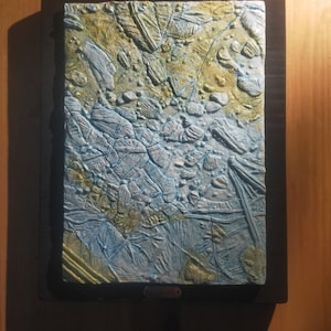 Pode incluir: Obra de arte abstrata texturizada em forma retangular, montada em uma moldura preta. A obra apresenta uma mistura de cores azul, amarelo e branco com uma superfície áspera e irregular. A peça é exibida em um fundo de madeira.