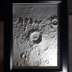 May include: A framed, three-dimensional relief map of the Copernicus crater on the Moon. The artwork is in shades of gray and white, with detailed textures and shadows. The black frame surrounds the detailed lunar surface. The artist's name and date are at the bottom.