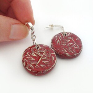 May include: A pair of red ceramic earrings with a white geometric pattern. The earrings are dangle style with silver hooks.