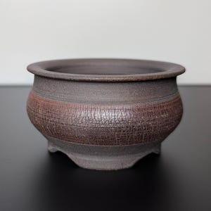 Op de afbeelding: Een donkerbruine keramische bonsai pot met een getextureerde, roodbruine band in het midden. De pot heeft een afgeronde vorm met een brede opening en drie kleine voetjes. Het algehele ontwerp is eenvoudig en rustiek.