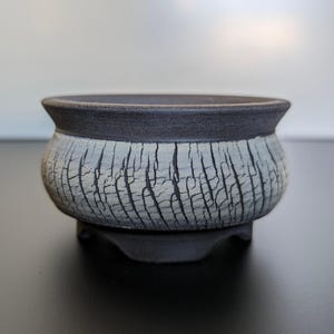 Op de afbeelding: Een kleine, ronde, zwart-witte keramische bonsai pot met een getextureerd, gebarsten glazuur. De pot heeft een brede, platte basis en een licht uitlopende rand.