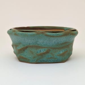 Può includere: Un vaso per bonsai in ceramica verde turchese con una superficie testurizzata. Il vaso ha una forma arrotondata con un bordo leggermente svasato.
