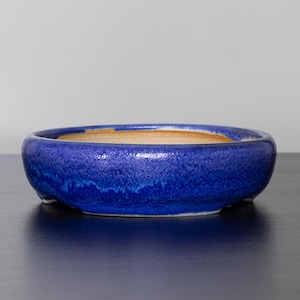 Puede incluir: Un cuenco de cerámica redondo y de perfil bajo con un vibrante esmalte azul. El cuenco tiene una superficie texturizada y un interior marrón claro. El borde es redondeado y el cuenco se apoya en tres pequeños pies. Este cuenco decorativo es adecuado para plantas o como acento decorativo.