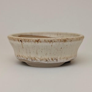 Op de afbeelding: Een kleine, gebroken witte keramische kom met een licht uitlopende rand en een afgeronde basis. De kom heeft een getextureerd oppervlak met bruine accenten langs de rand en de basis. De binnenkant is een donkerdere bruintint.
