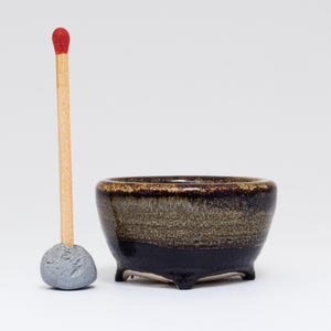 May include: A small, dark brown and grey ceramic bonsai pot with four feet. The pot is about the size of a matchstick.