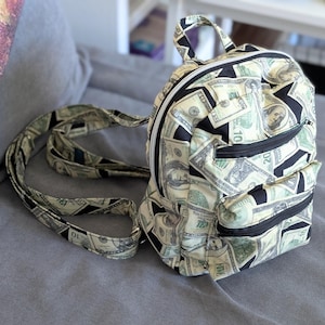 May include: A small backpack with a repeating pattern of US dollar bills. The bag has black zippers and trim, with a top handle and adjustable straps. The backpack is made of a fabric with a money print.