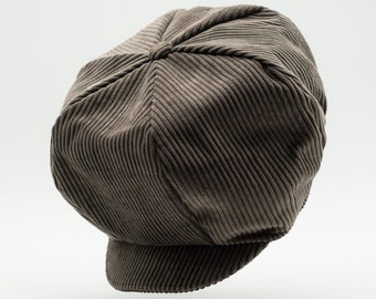 May include: A brown corduroy newsboy cap featuring a multi-paneled crown with a central button detail and a short, stiffened brim. The fabric has a distinct vertical ribbed texture throughout the entire surface of the rounded, structured hat.