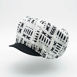 May include: A white cap featuring a repeating black geometric pattern of dots, lines, and chevron shapes. The cap has a solid black brim and a rounded, structured crown, providing a clear view of the fabric design and the contrast between the colors.