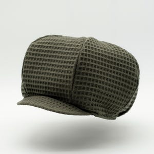 Könnte beinhalten: Eine olivgrüne Schiebermütze mit einer ausgeprägten Waffelstrickstruktur im gesamten Stoff. Die Mütze hat eine abgerundete, aus mehreren Paneelen bestehende Krone und einen kurzen, versteiften Schirm an der Vorderseite mit einem gleichmäßigen Gittermuster.